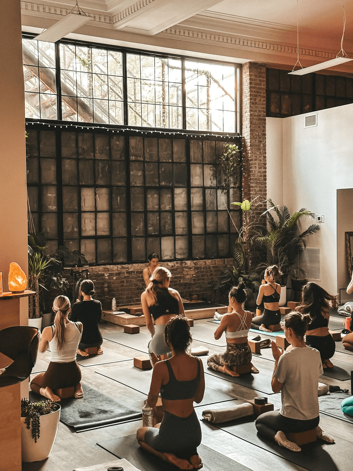 A circle of people sitting cross-legged, meditating with hands in a prayer position. Soft, warm lighting.